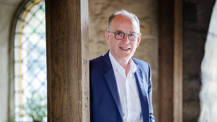 Ein Mann mit Brille, weißem Hemd und blauem Anzug steht in einem alten Klostergebäude und lehnt sich mit der Schulter gegen eine Holzsäule.