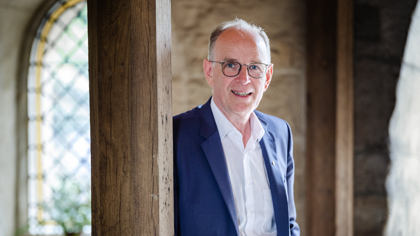 Ein Mann mit Brille, weißem Hemd und blauem Anzug steht in einem alten Klostergebäude und lehnt sich mit der Schulter gegen eine Holzsäule.